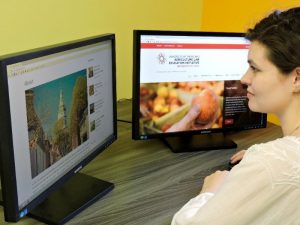 A person is seated at a desk using two computer monitors. The left monitor displays a webpage with an image of a building and text about its history. The right monitor shows the University of Maryland Agriculture Law Education Initiative website, featuring an image of a hand holding an apple. The setting is an office environment with a yellow wall.
