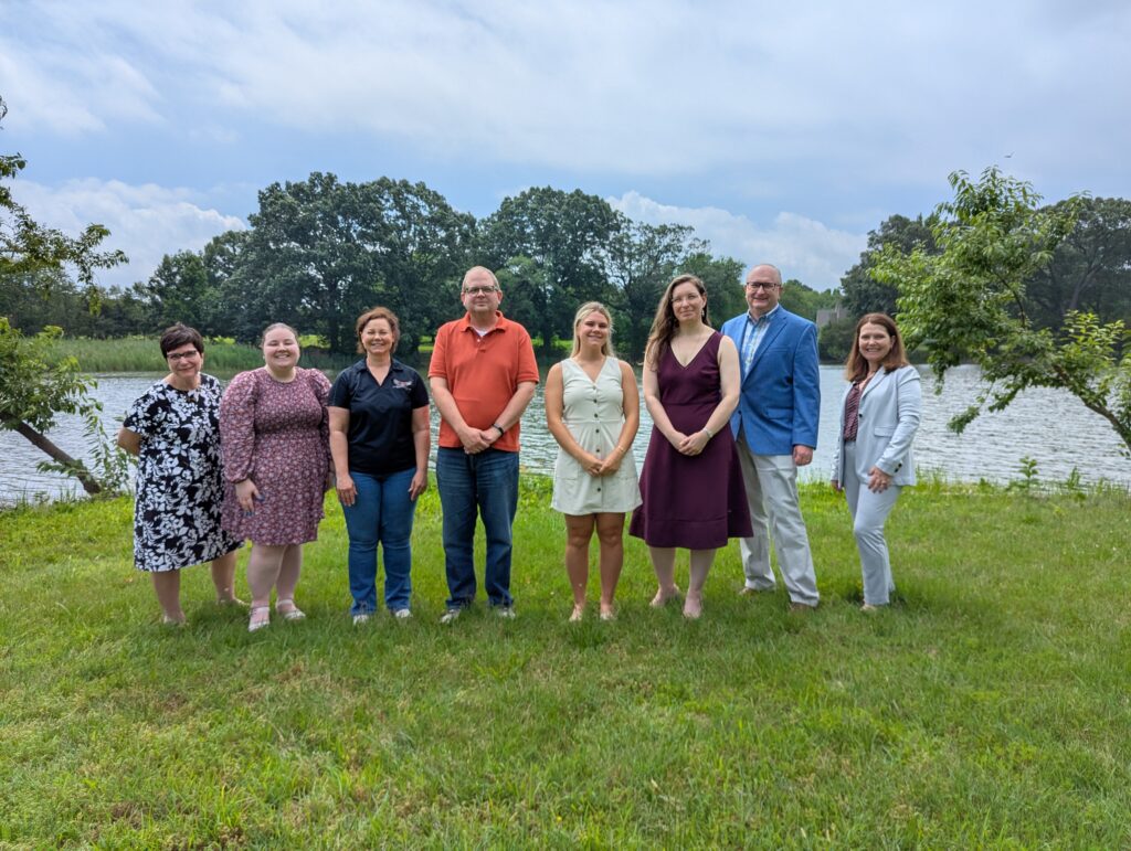 A group of people standing on a grassy area near a lake, with trees in the background on a cloudy day. The individuals are dressed in various casual and semi-formal attire.
