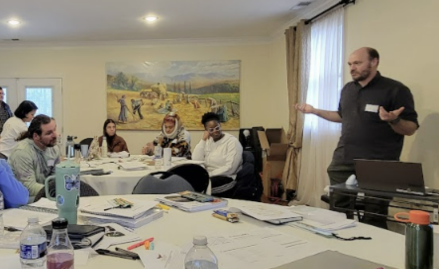 People sitting at a round table, listening to someone speak in the front of the room