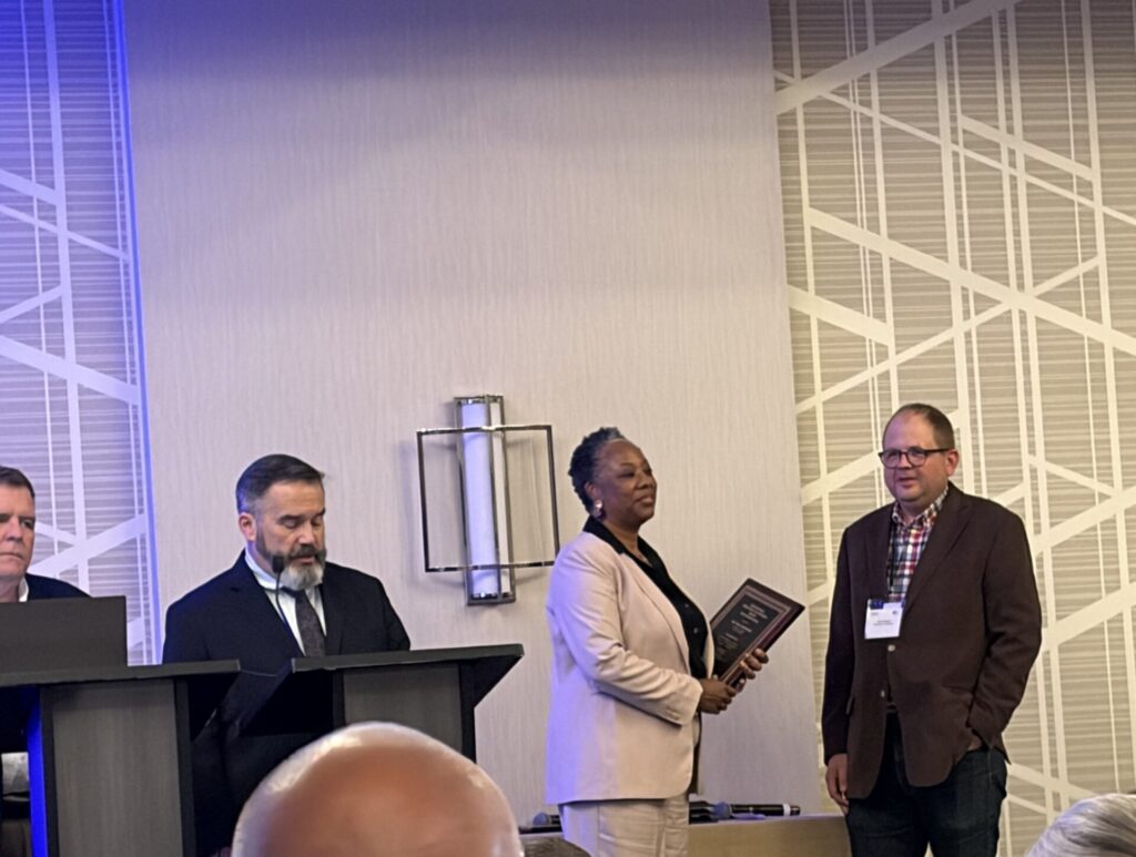 A woman in a light-colored suit presents a plaque to a man wearing a brown blazer at a conference event, while attendees watch from tables in the foreground. Two other people stand at podiums nearby.