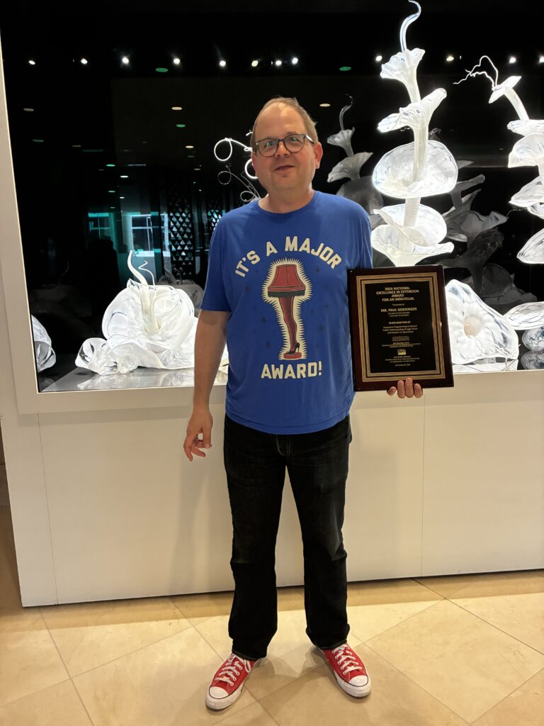 A man wearing glasses, a blue “It’s a Major Award!” T-shirt, black pants, and red sneakers holds a framed National Excellence in Extension award in front of a decorative glass art display.