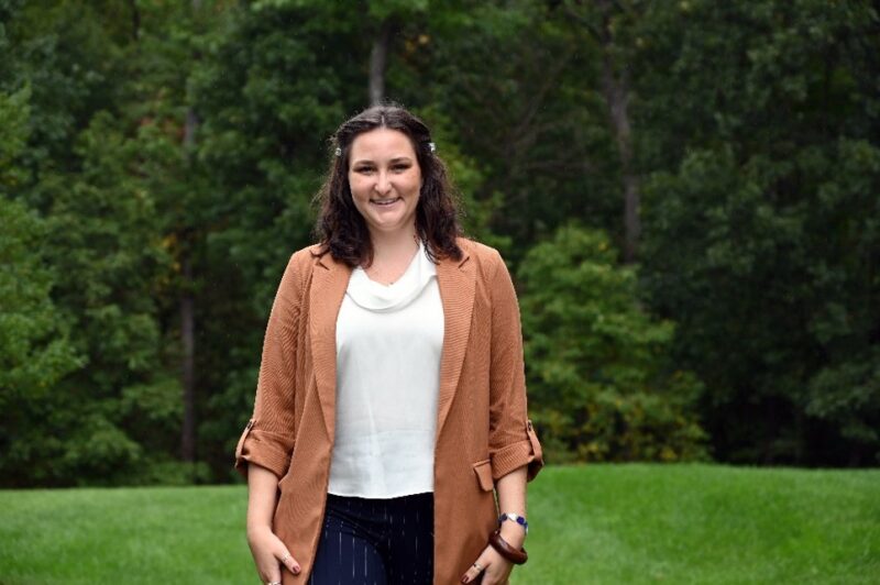 A woman smiling and standing outdoors in front of a wooded area. She is wearing a tan blazer over a white blouse and dark pants.