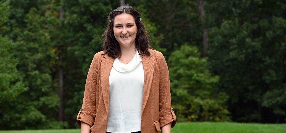 A woman smiling and standing outdoors in front of a wooded area. She is wearing a tan blazer over a white blouse and dark pants.