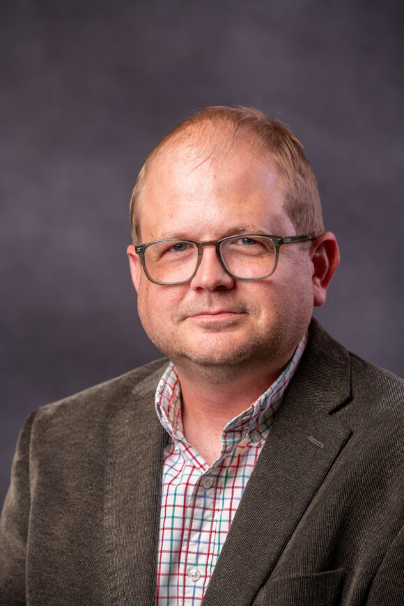 Professional headshot of a man in a brown blazer and a plaid shirt.