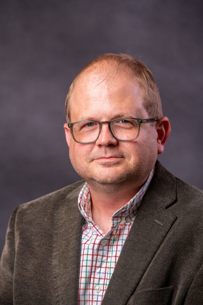 Professional headshot of a man in a brown blazer and a plaid shirt.